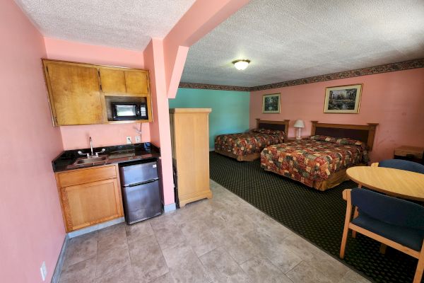 This image shows a hotel room with two double beds, a kitchenette with a fridge and microwave, a wooden wardrobe, and a small dining area with a round table.