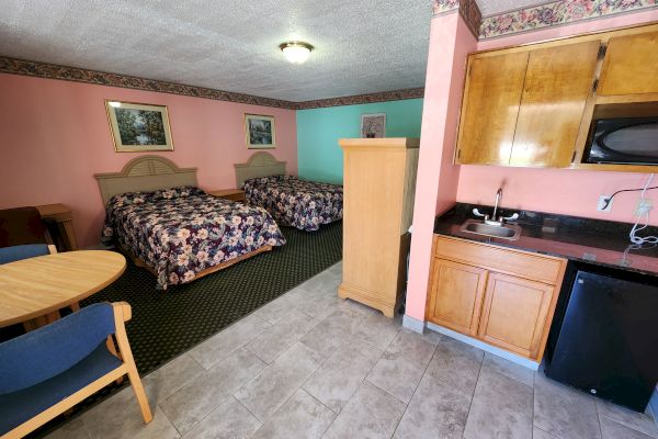 The image shows a hotel room with two beds, a small kitchen area, a round table with chairs, and wooden furniture. The walls are pastel-colored and decorated.