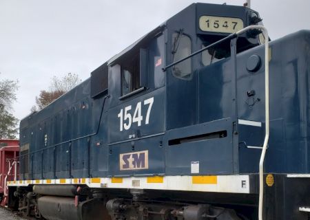 A blue train engine with the number "1547" on its side is seen on a track. The SM logo is present near the front of the locomotive.