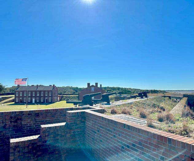 The image shows a historic fort with brick walls, an American flag, and buildings under a clear blue sky with sunlight.