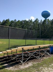 The image shows a baseball field with empty bleachers and a blue water tower in the background on a sunny day, ending the sentence.