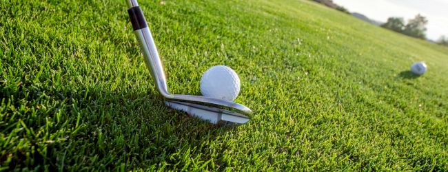 A close-up of a golf club preparing to hit a golf ball on a green grassy course, with another ball visible in the background.