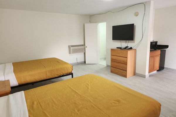 The image shows a hotel room with two beds, a TV on a dresser, a microwave, a fridge, and an open door to a bathroom.