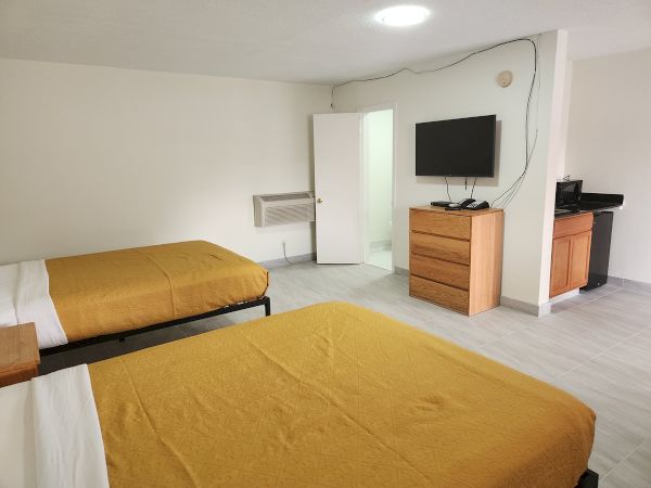 The image shows a hotel room with two beds, a TV on a dresser, a microwave, a fridge, and an open door to a bathroom.