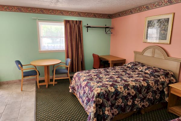 A hotel room with a bed, a table and chairs, a desk with a chair, hanging rack, window with curtain, and framed artwork on the wall.