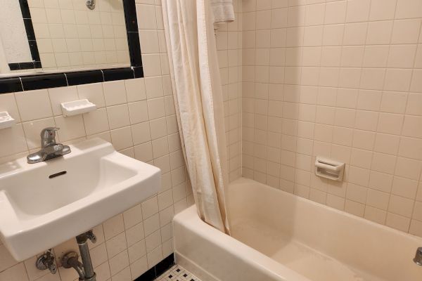 The image shows a bathroom with a white sink, a mirror above, a bathtub, and a shower curtain. The walls and tiles are white.