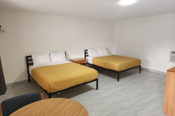 A hotel room with two double beds, white pillows, and mustard yellow covers, a wooden table, and a chair in the foreground.