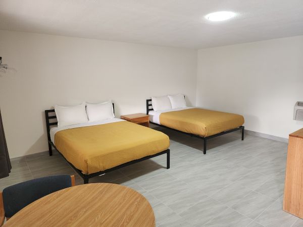 A hotel room with two double beds, white pillows, and mustard yellow covers, a wooden table, and a chair in the foreground.
