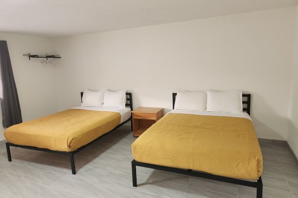 The image shows a room with two beds, yellow bedspreads, a wooden nightstand, gray curtains, and hangers on a wall rack.