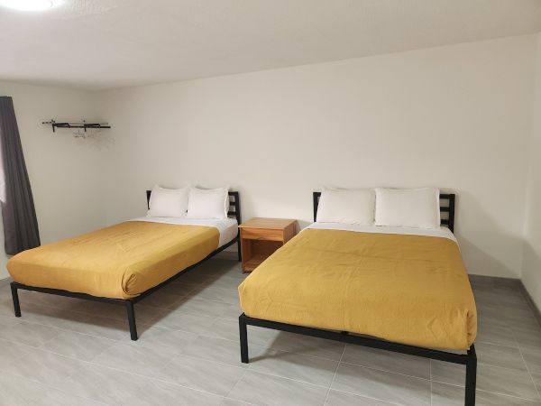 The image shows a room with two beds, yellow bedspreads, a wooden nightstand, gray curtains, and hangers on a wall rack.