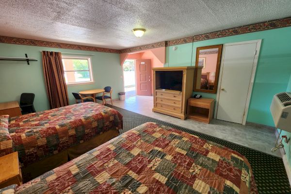 A hotel room with two beds, a TV on a wooden dresser, a small table with chairs, an air conditioner, and an open door to the outside.