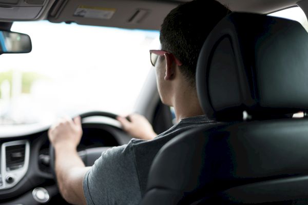 A person wearing sunglasses is driving a car, viewed from the back seat.