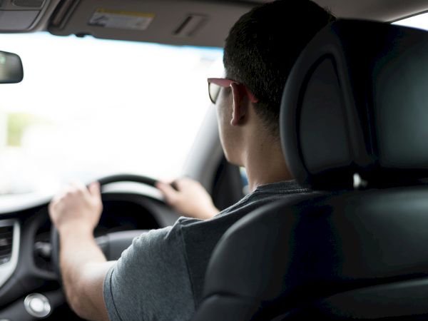 A person wearing sunglasses is driving a car, viewed from the back seat.