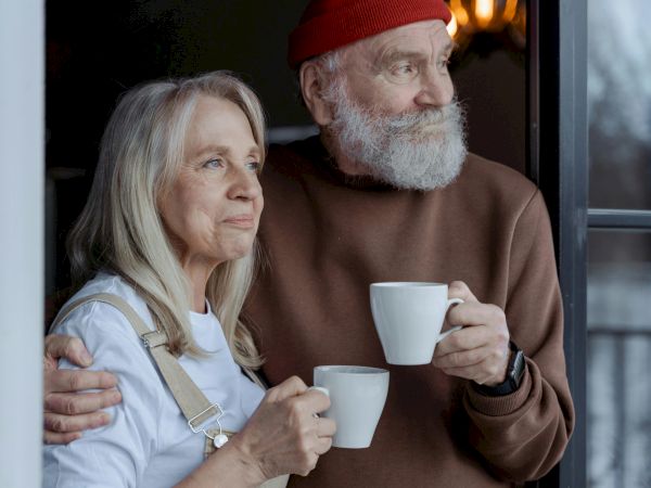 An elderly couple enjoys a warm drink while standing at a doorway, lovingly embracing each other as they look out thoughtfully.