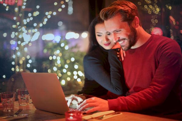 Two people are smiling and looking at a laptop while sitting at a cozy, decorated cafe with drinks and food on the table, surrounded by lights.