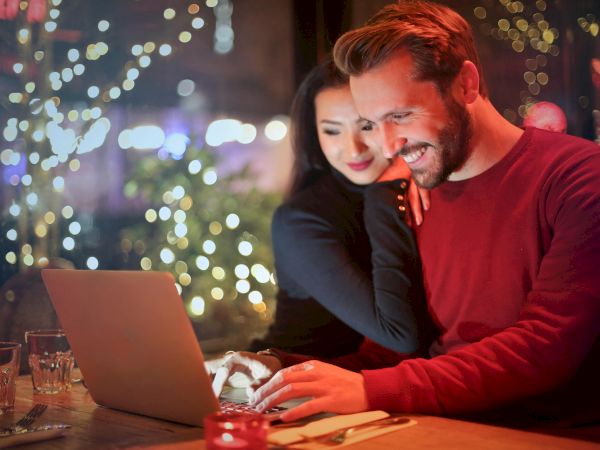 Two people are smiling and looking at a laptop while sitting at a cozy, decorated cafe with drinks and food on the table, surrounded by lights.