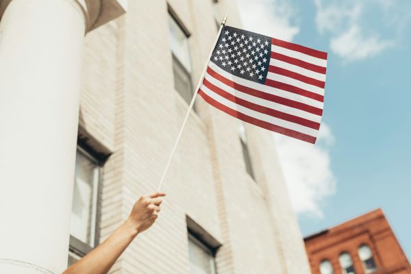 A hand is holding an American flag in front of a light-colored building with windows, with a blue sky and another building in the background.