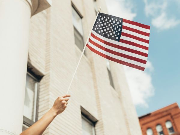 A hand is holding an American flag in front of a light-colored building with windows, with a blue sky and another building in the background.