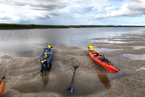 Two kayaks, one blue and one orange, rest on a sandy riverbank with two paddles. Marshy landscape and cloudy skies are visible in the background.