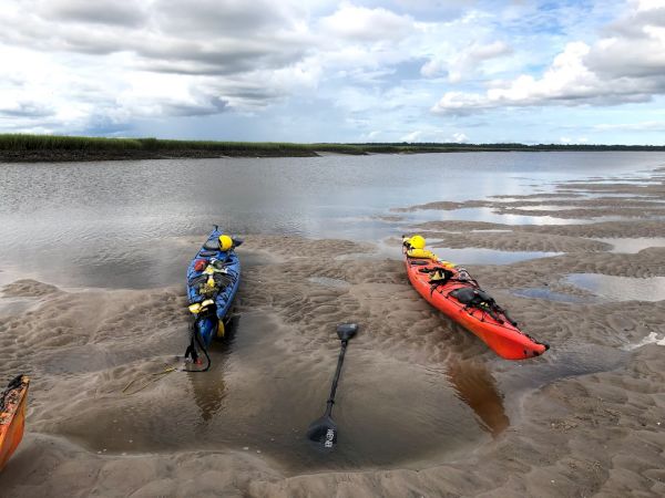Two kayaks, one blue and one orange, rest on a sandy riverbank with two paddles. Marshy landscape and cloudy skies are visible in the background.