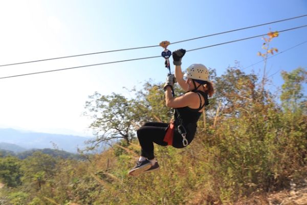 A person wearing a helmet and harness is zip lining over a scenic outdoor landscape with trees and hills in the background, enjoying the adventure.