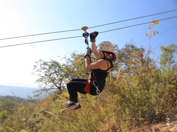 A person wearing a helmet and harness is zip lining over a scenic outdoor landscape with trees and hills in the background, enjoying the adventure.