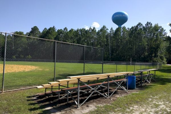 The image shows an empty set of bleachers next to a baseball field with a water tower and trees in the background.