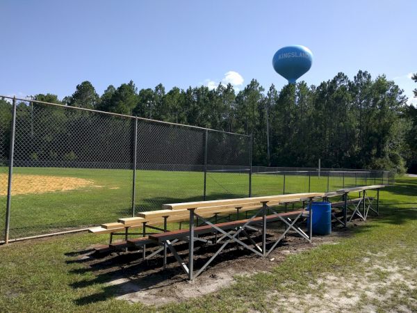 The image shows an empty set of bleachers next to a baseball field with a water tower and trees in the background.