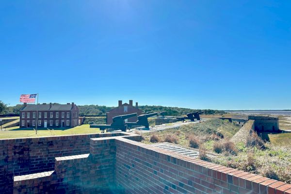 A fort with brick walls, historical buildings, and an American flag under a clear blue sky. There's a grassy area and pathways visible.