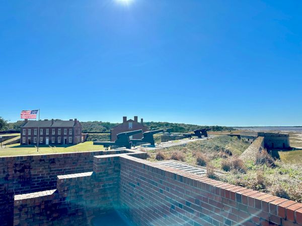 A fort with brick walls, historical buildings, and an American flag under a clear blue sky. There's a grassy area and pathways visible.