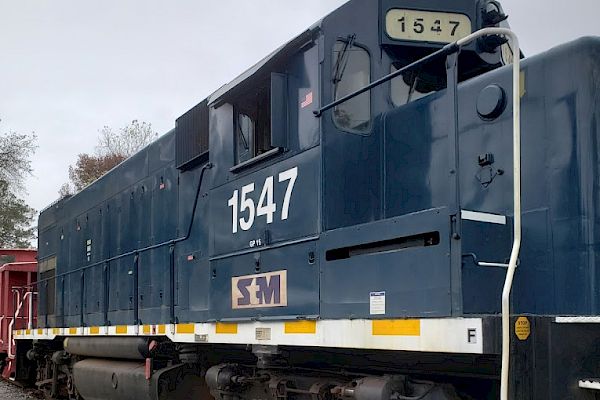 The image shows a blue locomotive engine with the number 1547 on the side, parked on a railway track with some trees visible in the background.