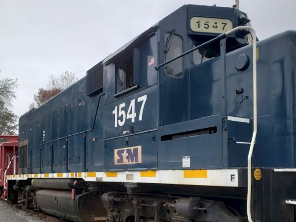 The image shows a blue locomotive engine with the number 1547 on the side, parked on a railway track with some trees visible in the background.
