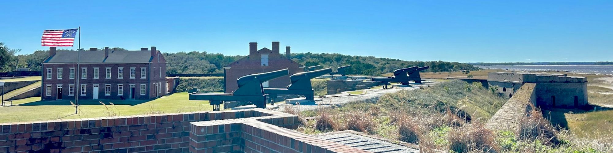 A historic fort with brick structures, green grassy areas, and an American flag on a pole, against a clear blue sky and horizon.