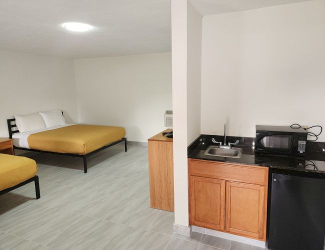 The image shows a motel room with two beds, a kitchenette with a sink, microwave, and mini-fridge, and basic furnishings.