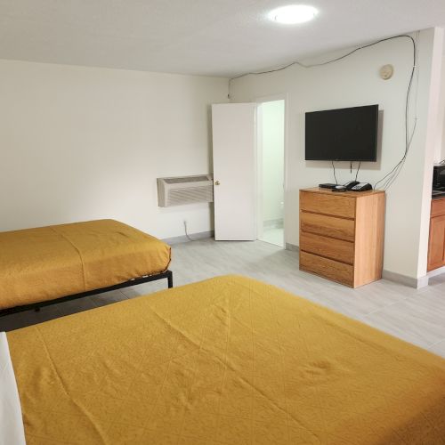 The image shows a hotel room with two beds, a TV, a wooden dresser, and a small kitchenette area.