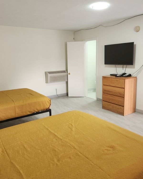 The image shows a hotel room with two beds, a TV, a wooden dresser, and a small kitchenette area.