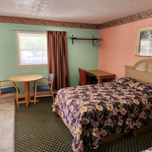 A hotel room with a bed, a round table with two chairs, a desk with a chair, and a window with a curtain, featuring colorful walls and bedding.