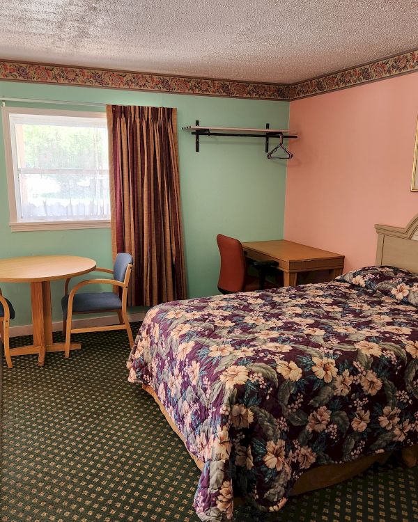 A hotel room with a bed, a round table with two chairs, a desk with a chair, and a window with a curtain, featuring colorful walls and bedding.