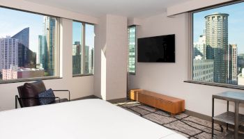 A modern hotel room with large windows offers a cityscape view, featuring a bed, armchair, wall-mounted TV, and wooden console.