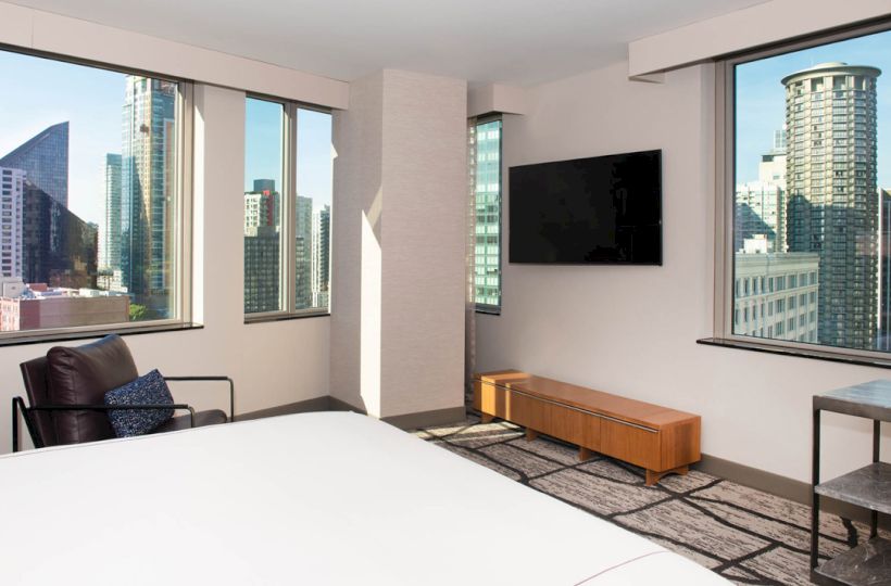 A modern hotel room with large windows offers a cityscape view, featuring a bed, armchair, wall-mounted TV, and wooden console.