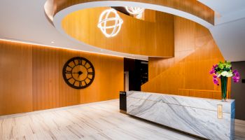 A modern lobby with a white marble reception desk, a large wall clock, wooden accents, spiral staircase, and decorative ceiling lights.