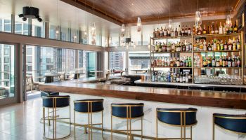 A modern bar with a sleek, wooden countertop, comfortable blue and gold bar stools, and shelves stocked with various bottles of alcohol.