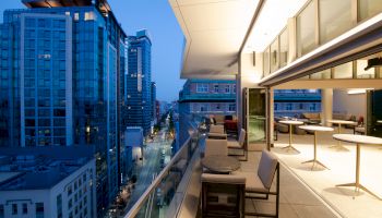 A modern cityscape taken from a balcony featuring outdoor seating, high-rise buildings, and a street view at dusk.