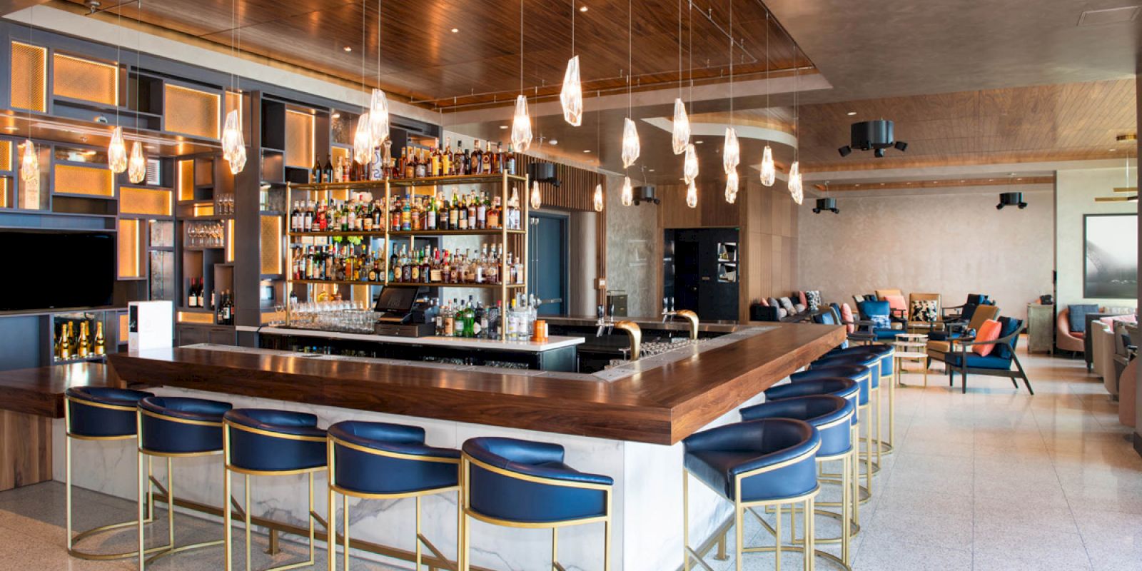 The image shows a modern bar with blue and gold stools, a well-stocked liquor shelf, pendant lights, and a spacious seating area in the background.