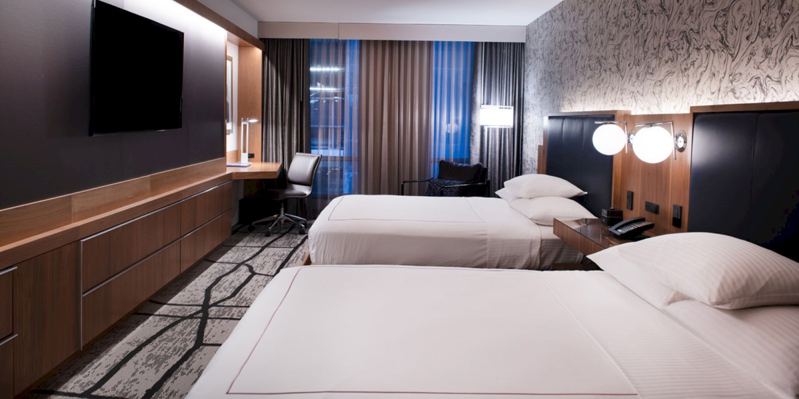 A modern hotel room with two double beds, a flat-screen TV, a desk and chair near a window, contemporary decor, and dim lighting.