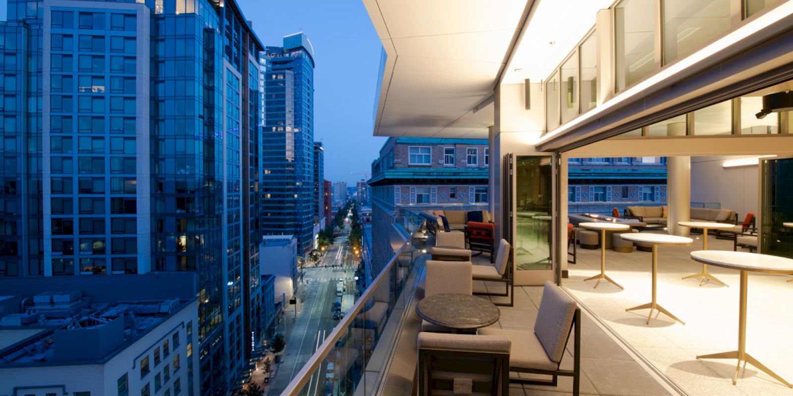 A modern cityscape showing tall buildings at dusk, with a view from a balcony that has tables and chairs. Lights are on in buildings and street.