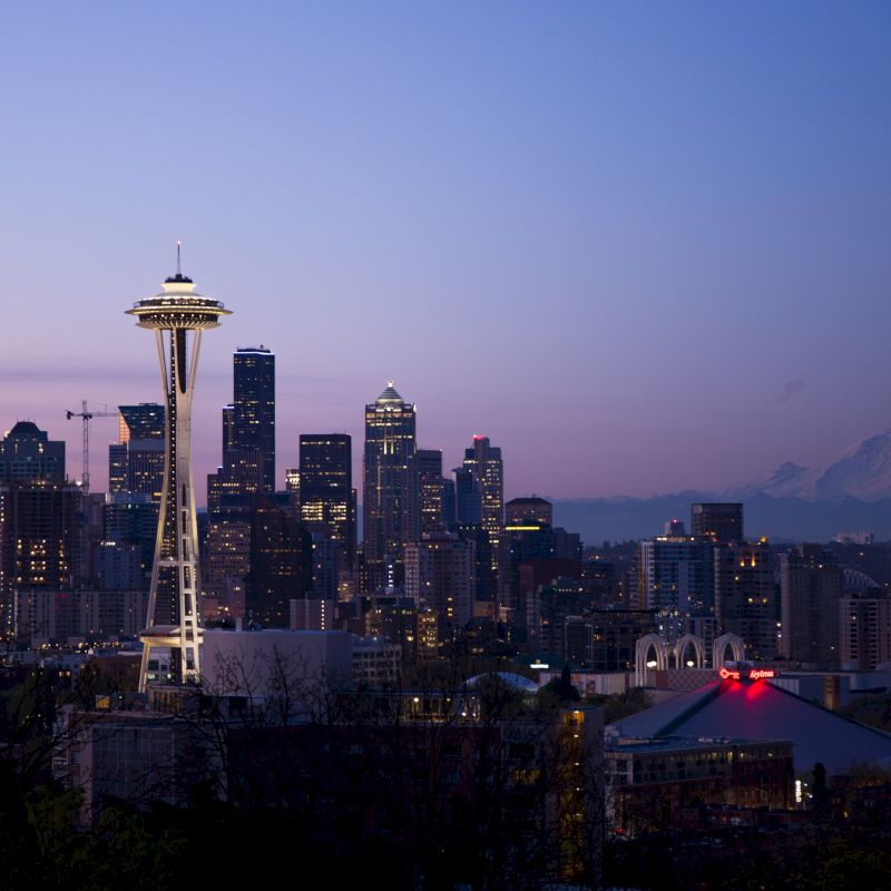 This is a cityscape of Seattle at dusk, featuring the iconic Space Needle, surrounding skyscrapers, and distant Mount Rainier under a twilight sky.