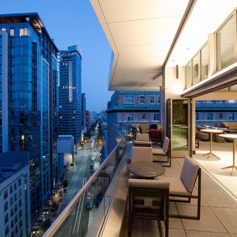 A modern urban balcony at dusk with city views, outdoor seating, and an indoor area with tables and chairs visible through open glass doors.