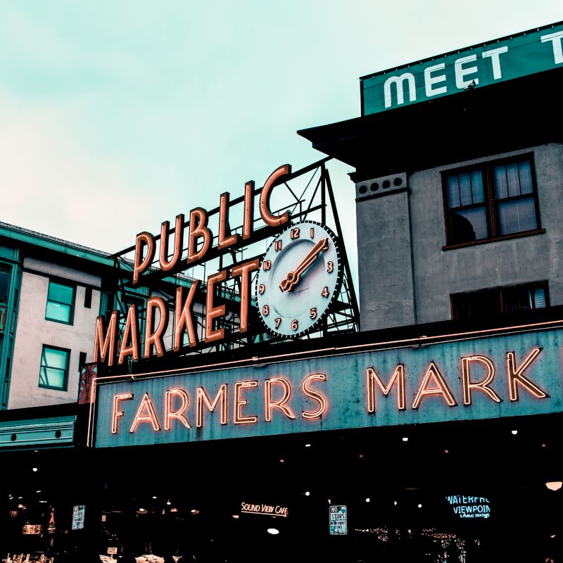 A neon sign reads "Public Market" with a clock beneath it, and a larger neon sign below reads "Farmers Market" against a backdrop of buildings.