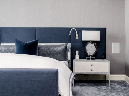 A modern bedroom featuring a navy blue upholstered bed, white and gray bedding, a nightstand with a lamp, and a decorative object on the nightstand.